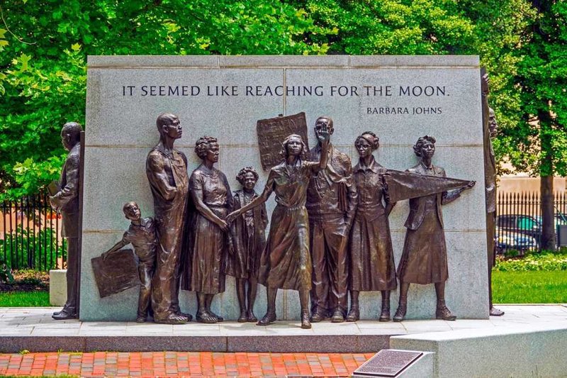 The image is of the Virginia Civil Rights Memorial which includes statues of adults and children surrounding a large grey block. The side of the memorial that is featured here includes a quote that says, "It seemed like reaching for the moon." -Barbara Johns