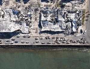 Aerial view of fire-damaged buildings and burnt vehicles along a coastal road.