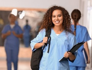 a nursing student with a backpack and notebook smiles while fellow students talk and walk down the hospital hallway in the background