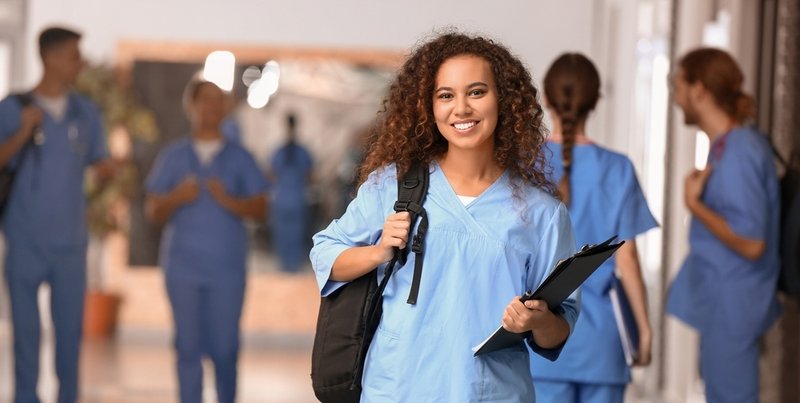 a nursing student with a backpack and notebook smiles while fellow students talk and walk down the hospital hallway in the background