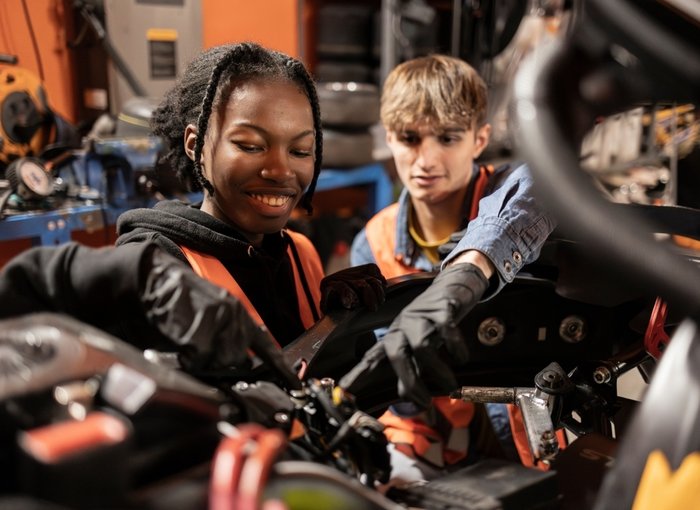 Two young apprentices wearing safety vests and gloves work together on in a workshop, focusing closely on mechanical components.