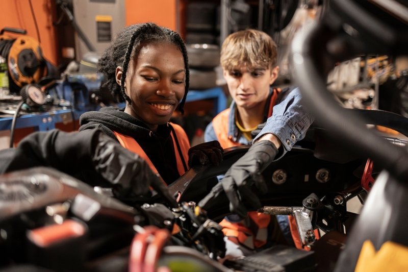 Two young apprentices wearing safety vests and gloves work together on in a workshop, focusing closely on mechanical components.
