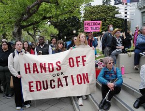 The image shows a group of people protesting with a large white sign with red lettering that reads "Hands Off Our Education."