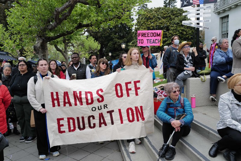 The image shows a group of people protesting with a large white sign with red lettering that reads "Hands Off Our Education."
