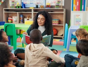 A preschool teacher reads a book to students while they sit on the floor.