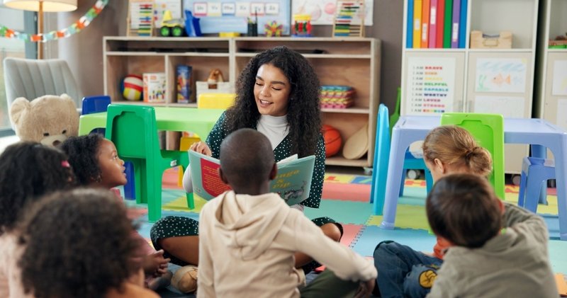 A preschool teacher reads a book to students while they sit on the floor.